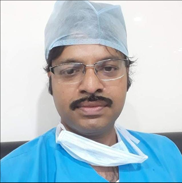 Portrait of Dr. Sujit Gharai wearing surgical scrubs, cap, and glasses, looking at the camera inside the eye clinic