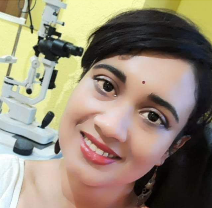 Close-up portrait of a smiling female eye specialist inside an eye clinic, with a slit-lamp microscope blurred in the background